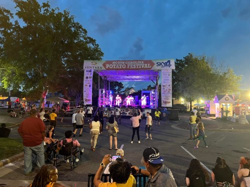 A lively outdoor concert at the North Carolina Potato Festival, with a crowd enjoying the performance under a twilight sky.