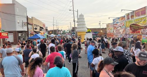 A crowded street fair with food stalls, people walking, and a large ice cream cone structure in the background.