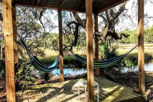 Two hammocks hang under a wooden gazebo, surrounded by trees and a serene water view.