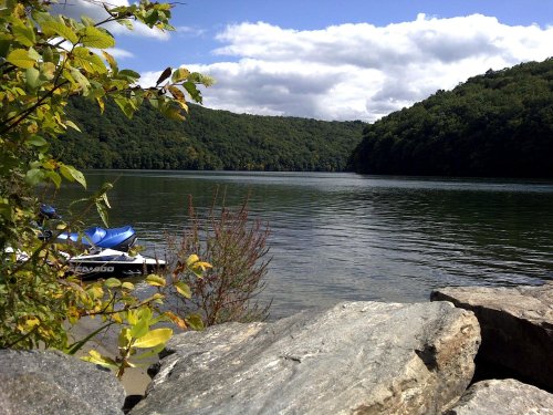 A serene lake surrounded by green hills, with a rocky shore and a blue boat moored nearby under a partly cloudy sky.