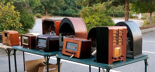 A row of vintage radios displayed on tables outdoors, surrounded by trees and an empty parking lot.