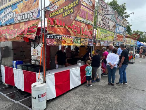 A busy food stall area at a fair, featuring various vendors selling turkey legs and other snacks. People are in line.
