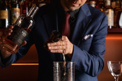 A bartender in a suit pours a spirit from a bottle into a jigger over a glass, with a bar backdrop.