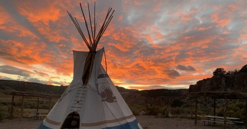 A teepee stands against a vibrant sunset sky with colorful clouds and a mountainous landscape in the background.