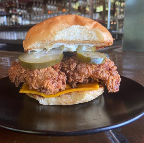A crispy fried chicken sandwich with pickles and cheese on a soft bun, served on a black plate.