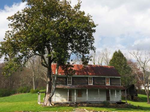 A weathered two-story house with a red roof, surrounded by trees and grassy fields under a partly cloudy sky.