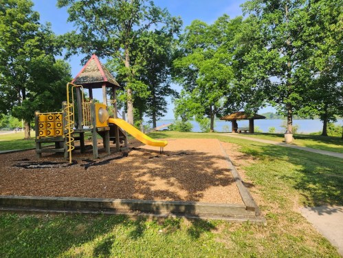 A playground with a slide and climbing structure, surrounded by trees and a view of a lake in the background.