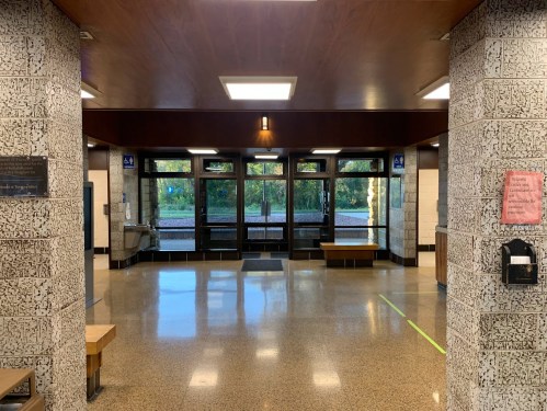 Interior view of a spacious lobby with large windows, benches, and a water fountain, featuring textured walls and polished floors.