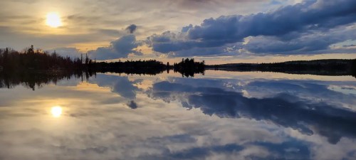 A serene lake at sunset, reflecting clouds and the sun, surrounded by trees and a peaceful landscape.
