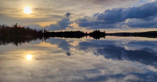 A serene lake at sunset, reflecting clouds and the sun in calm waters, surrounded by trees.