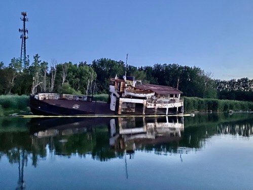 Abandoned boat partially submerged in calm water, surrounded by trees and a cell tower in the background at dusk.