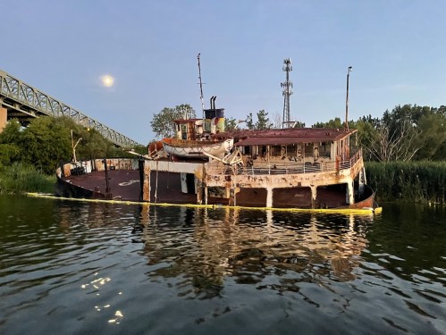 An abandoned, rusted boat partially submerged in water, surrounded by greenery, with a bridge and moon in the background.