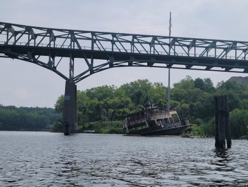 A partially submerged boat near a bridge, surrounded by greenery and calm water on a sunny day.