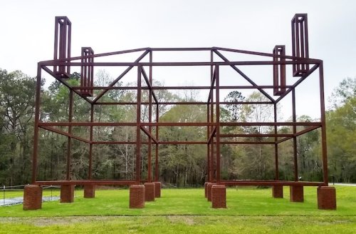A large, rust-colored steel frame structure stands on green grass, surrounded by trees under a cloudy sky.