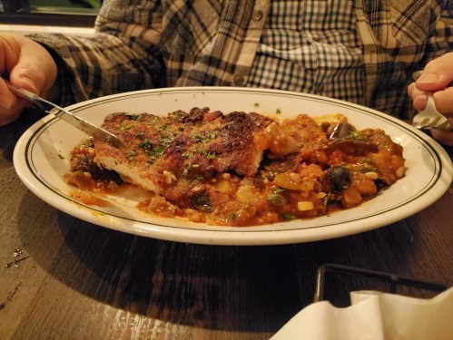 A plate of seasoned meat with a rich vegetable sauce, served on a wooden table, with a hand holding a fork and knife.