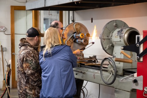 Three individuals observe a metalworking process involving flames and machinery in a workshop setting.