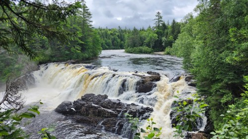A scenic view of a waterfall cascading over rocky ledges, surrounded by lush green trees and a cloudy sky.
