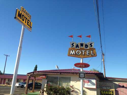 Signage for Sands Motel with retro design, flags, and a "Vacancy" notice under a clear blue sky.