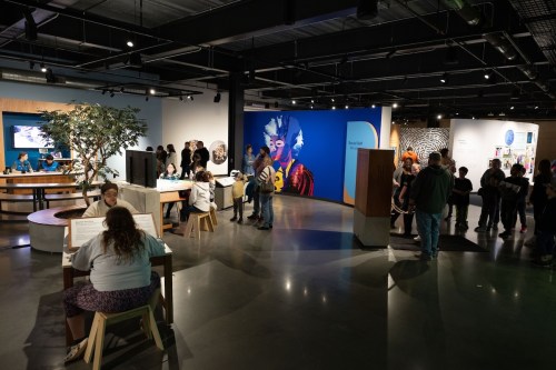 A vibrant museum space with visitors exploring exhibits, seated at tables, and a colorful mural on the wall.
