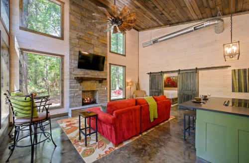 Cozy living room with a red sofa, stone fireplace, large windows, and rustic decor in a forested setting.
