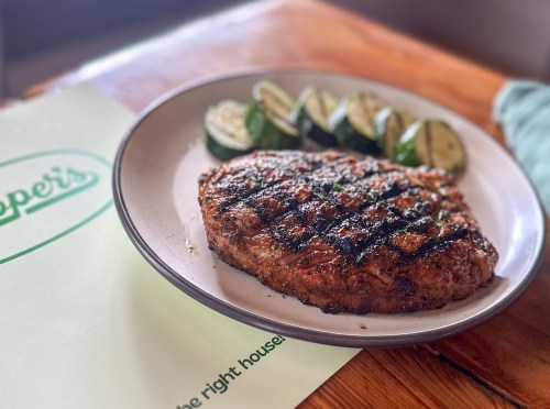 A grilled steak on a plate, accompanied by sliced zucchini, on a wooden table with a menu in the background.
