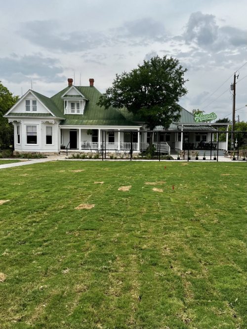 A large white house with a green roof, surrounded by a grassy lawn and trees, featuring a porch and a sign.