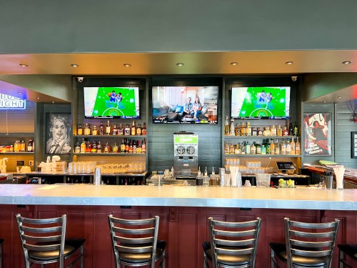 A bar with a marble countertop, two TVs showing sports, and shelves stocked with various drinks and glassware.