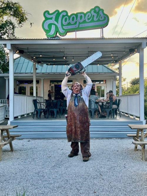 A person in a bloodied apron holds a chainsaw aloft in front of a restaurant named Hooper's.
