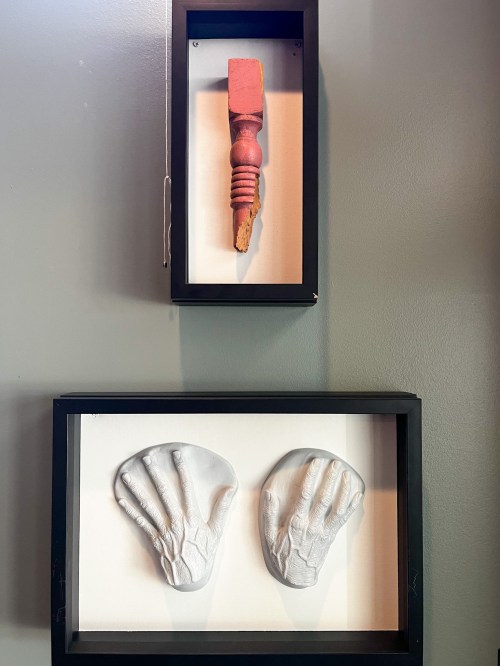 A wall display featuring a wooden object above two white hand sculptures in black frames.