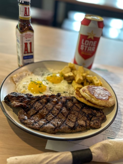 A plate with a grilled steak, two eggs, hash browns, a pancake, a bottle of A1 sauce, and a can of Lone Star beer.