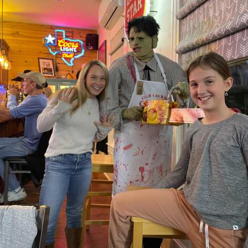 A woman and a girl pose with a zombie mannequin holding a tray, in a lively bar setting with neon lights.