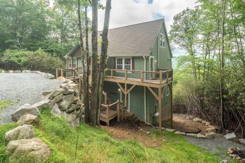 A green two-story house on stilts surrounded by trees and gravel, with a wooden deck and stone pathway.