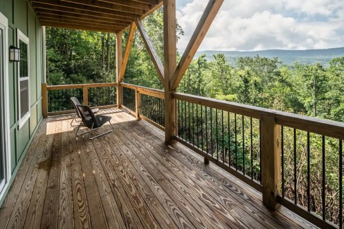 A wooden deck with a chair overlooks a lush green forest and distant mountains under a cloudy sky.