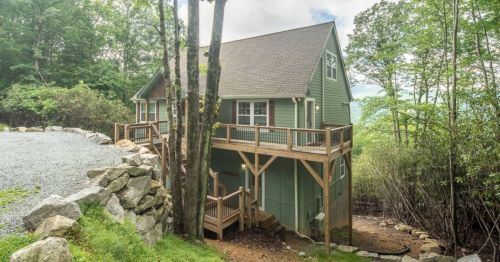 A green house with a wooden deck, surrounded by trees and rocky terrain, set in a serene outdoor environment.