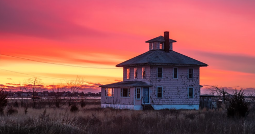 A weathered house stands in a field against a vibrant sunset sky with shades of pink and orange.
