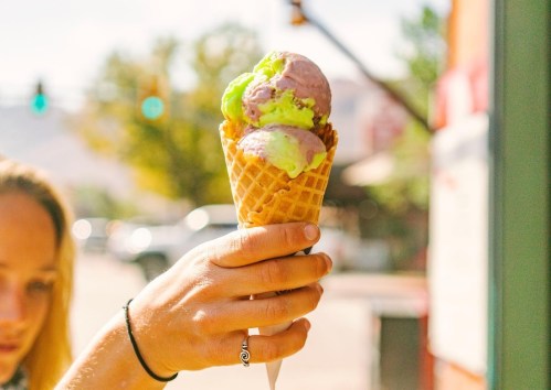 A hand holds a waffle cone with three scoops of colorful ice cream: purple, green, and white, against a sunny background.
