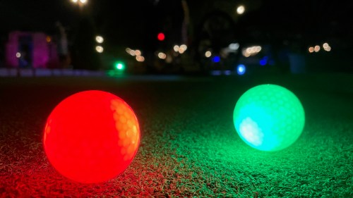 Two glowing golf balls, one red and one green, sit on a grassy surface at night, with colorful lights in the background.