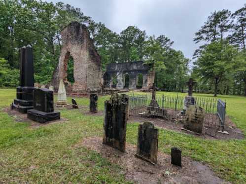 A cemetery with weathered gravestones and the ruins of a brick structure surrounded by trees under a cloudy sky.