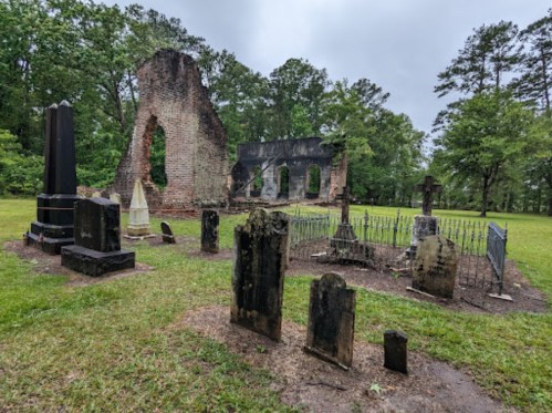 A cemetery with weathered gravestones and the ruins of a brick structure surrounded by trees under a cloudy sky.