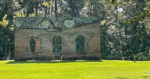 Ruins of a brick structure with arched windows, surrounded by green grass and trees in a wooded area.