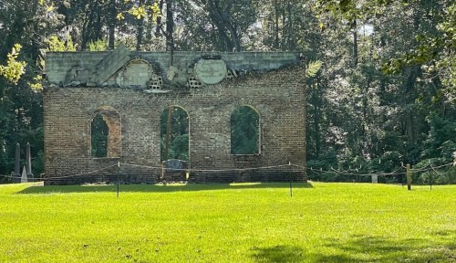 Ruins of a brick structure with arched windows, surrounded by green grass and trees in a serene outdoor setting.