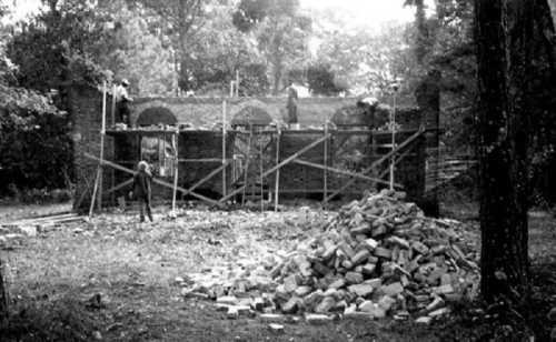 Black and white image of workers constructing a brick structure with scaffolding and a pile of bricks nearby.