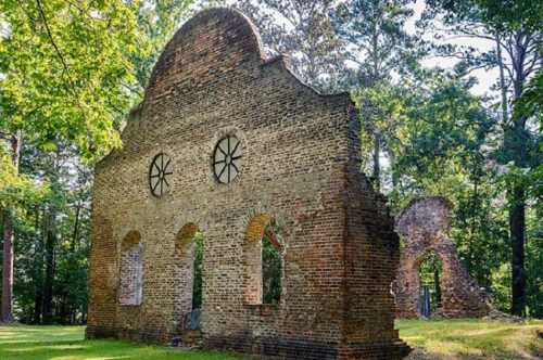 Ruins of a brick structure with arched windows, surrounded by lush green trees in a serene outdoor setting.