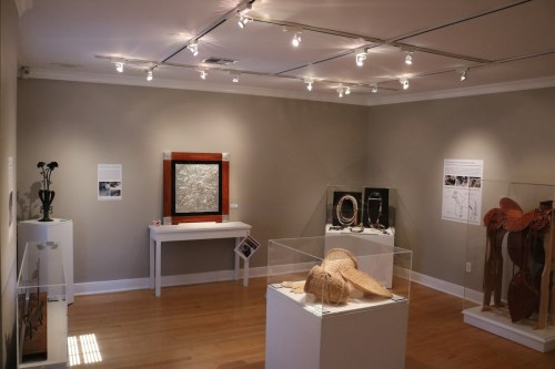 Art gallery interior featuring various exhibits, including sculptures and a framed artwork, under bright lighting.