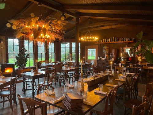 Cozy restaurant interior with wooden tables, candles, and a warm chandelier, surrounded by large windows and greenery.