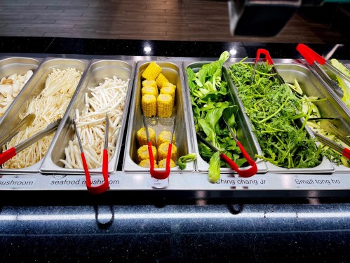 A buffet display with various fresh vegetables, including corn, mushrooms, and leafy greens, in metal trays.