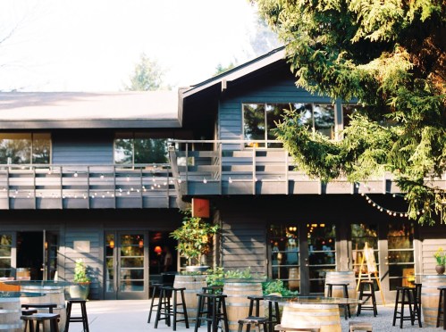 A cozy wooden building with large windows, surrounded by trees and outdoor seating with barrels and chairs.
