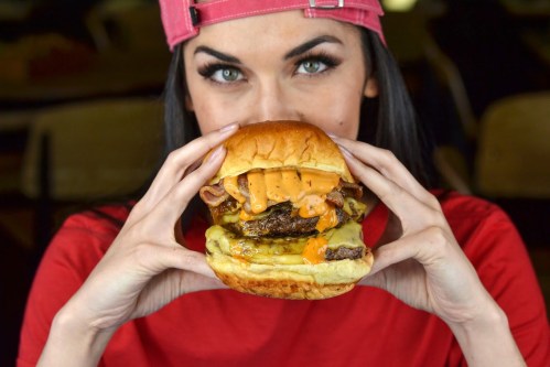 A person in a red shirt and pink cap holds a large cheeseburger with melted cheese and toppings, ready to take a bite.