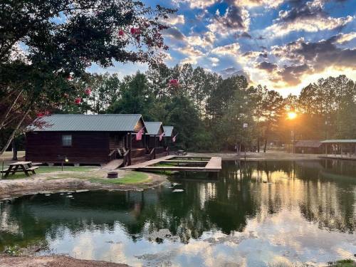 A serene lakeside scene with wooden cabins, a picnic table, and a sunset reflecting on the water.