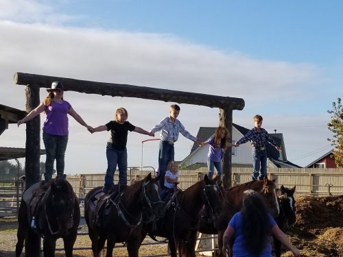 Five children stand on horseback, holding hands, with a barn and cloudy sky in the background.